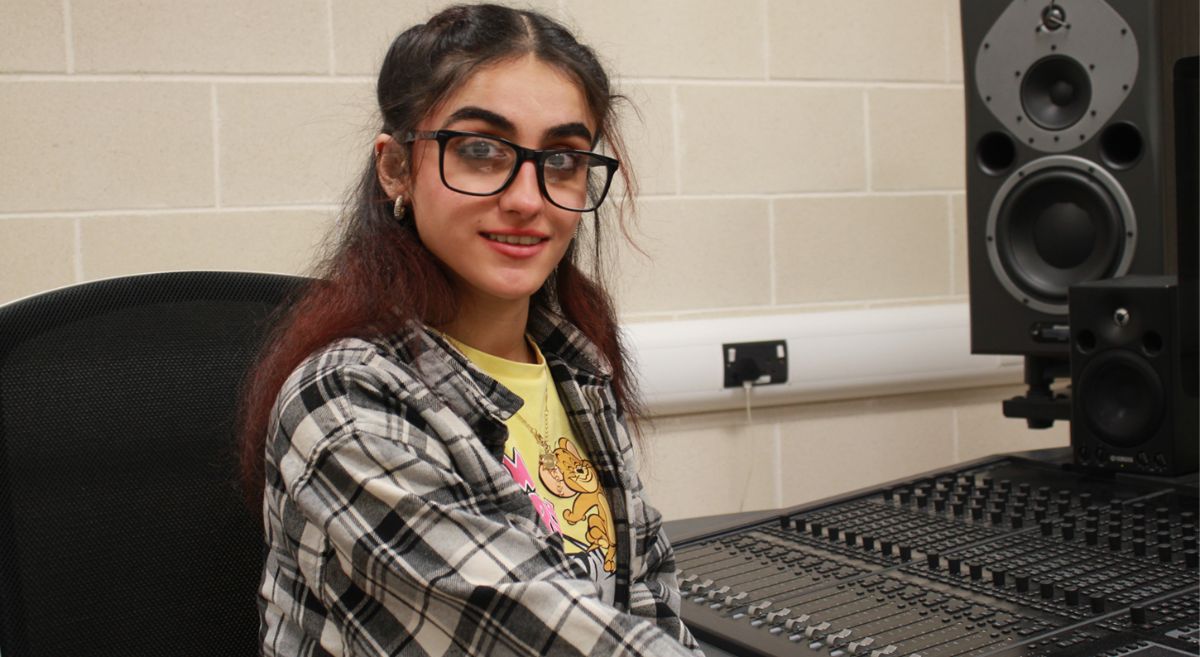 Female wearing a checked shirt and glasses sitting beside a sound desk and speaker.